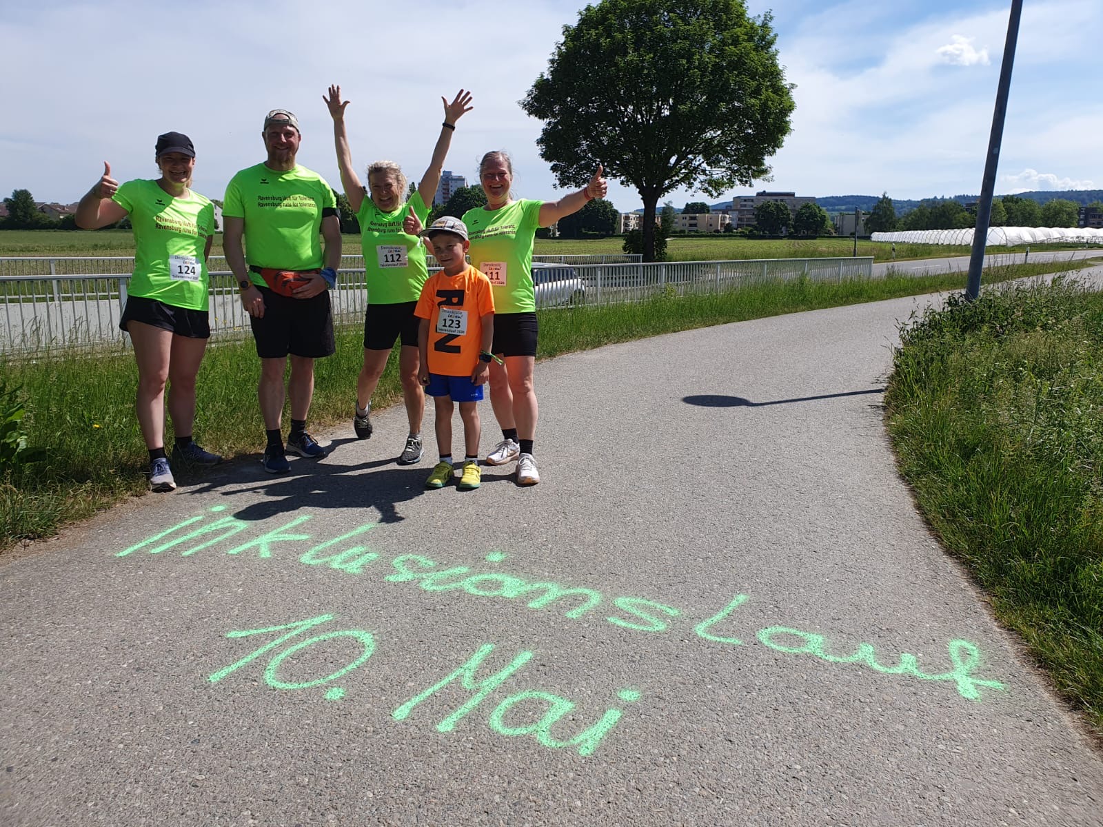 Personen in neongelbem Lauftrikots schaut fröhlich in die Kamera. Vor der Gruppe kann man auf der Straße lesen: "Inklusionslauf 10. Mai"