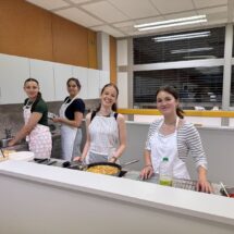 Drei Schülerinnen bereiten spanische Delikatessen zu und blicken lachend in die Kamera.