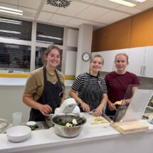Drei Schülerinnen bereiten spanische Delikatessen zu und blicken lachend in die Kamera.